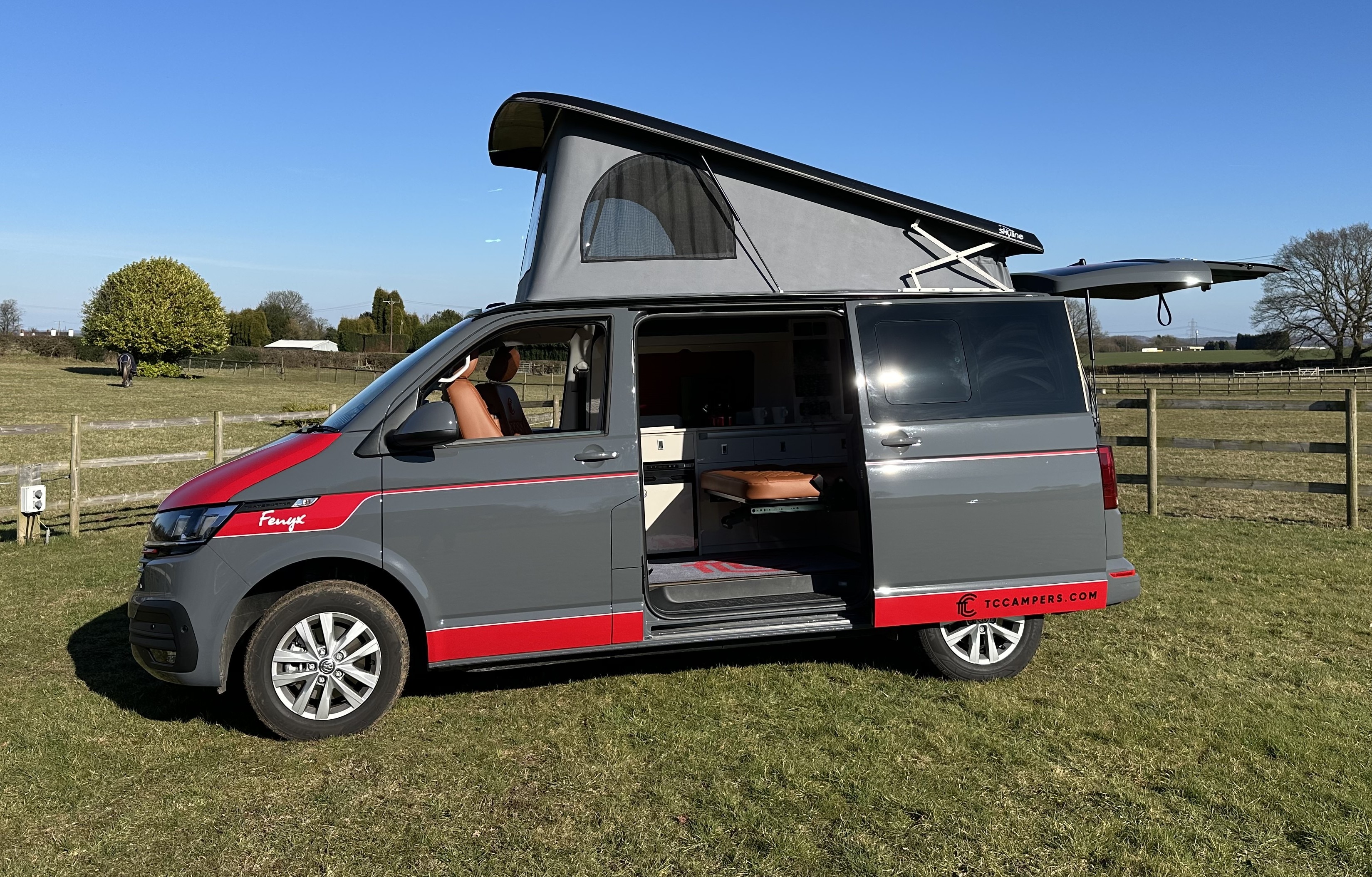 Fenyx, a grey VW Transporter campervan with red detailing, the pop-top roof up and the side door open to the interior, parked on grass