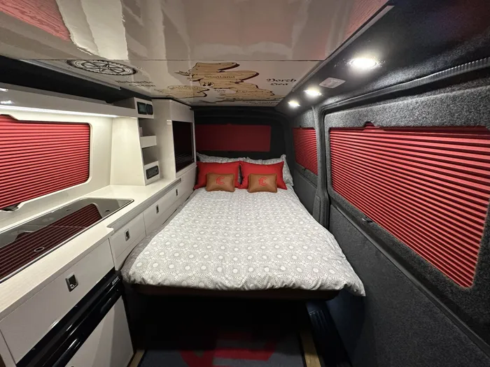 Fenyx with the bed made up, red blackout blinds down and the kitchen alongside